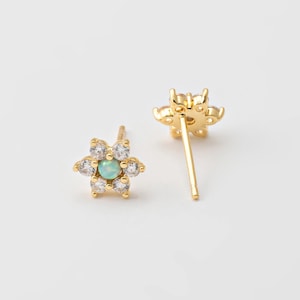May include: A pair of gold-colored flower stud earrings. Each earring features a central light blue gemstone surrounded by clear, round gemstones. The earrings are set against a white background.
