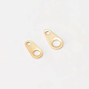 Puede incluir: Dos pequeños tiradores de cremallera de metal dorado. Cada tirador tiene forma de lágrima con un agujero circular en la parte inferior y un agujero más pequeño en la parte superior. Los tiradores están sobre un fondo blanco.