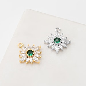Könnte beinhalten: Zwei blütenförmige Anhänger. Einer ist goldfarben mit einem grünen Mittelstein und weißen Akzenten. Der andere ist silberfarben mit einem grünen Mittelstein und weißen Akzenten. Beide haben eine Schlaufe zum Befestigen an einer Kette oder anderem Schmuck.