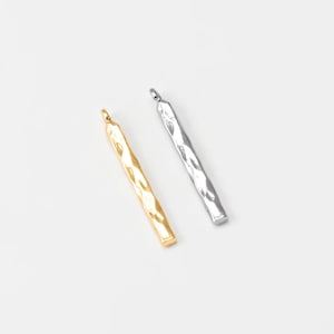 Puede incluir: Dos colgantes de metal texturizado. Uno es de color dorado, el otro plateado. Ambos son rectangulares con un acabado martillado y un pequeño lazo en la parte superior para colgar. Los colgantes se muestran sobre un fondo blanco.