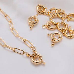 Może przedstawiać: Złote elementy biżuterii, w tym naszyjnik z wydłużonymi ogniwami i kilka zapięć typu toggle. Zapięcia są okrągłe z małym prętem i zamknięciem kulkowym. Elementy biżuterii są prezentowane na białej powierzchni.