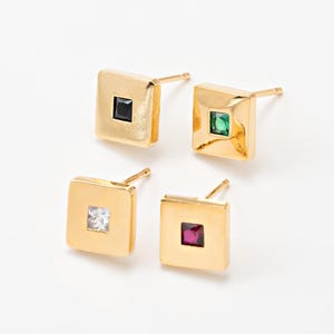 May include: Four gold-colored square stud earrings, each with a different colored gemstone: black, green, clear, and red. The earrings are displayed on a white surface.
