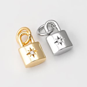 Może przedstawiać: Dwa zawieszki w kształcie kłódki, jeden złoty, a drugi srebrny, z małym diamentem w środku każdej kłódki. Diament jest osadzony w kształcie gwiazdy.