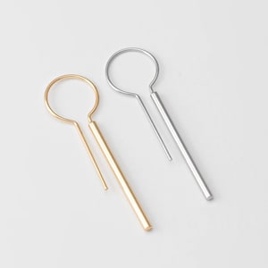 Op de afbeelding: Een paar minimalistische oorbellen, een gouden en een zilveren, met een eenvoudige cirkel en een lange, rechte staaf.