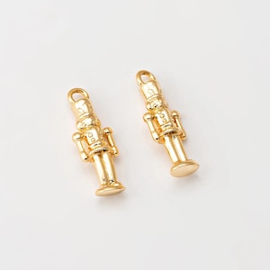 Puede incluir: Dos colgantes de metal dorado con forma de soldados cascanueces. Cada colgante tiene un lazo en la parte superior para sujetarlo a un collar o pulsera. Los colgantes están sobre un fondo blanco.