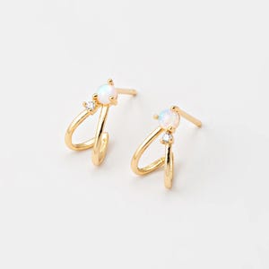 May include: A pair of gold-colored hoop earrings. Each earring features a double hoop design with a small, round, white opal stone and a tiny clear crystal accent. The earrings are set against a white background.