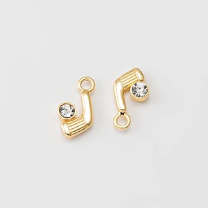 May include: Two gold-colored golf club charms with a clear crystal accent. The charms are detailed with a textured club head and a small loop for attaching to jewelry. The golf club charms are set against a white background.