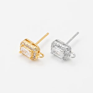 Puede incluir: Dos pendientes de botón, uno dorado y otro plateado, cada uno con un centro rectangular de piedra preciosa transparente rodeado de piedras más pequeñas y transparentes. Los pendientes tienen un poste y un pequeño lazo en la parte inferior.