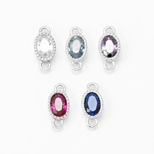 Può includere: Cinque ciondoli ovali con pietre preziose in vari colori: trasparente, blu, viola, rosso e blu scuro. Ogni ciondolo è incastonato in una montatura in metallo argentato con un bordo perlato e un anello ad ogni estremità.