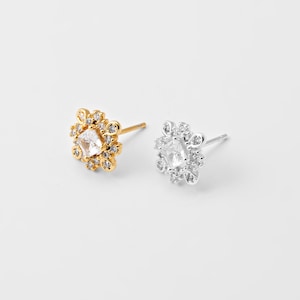 May include: Two stud earrings, one gold-toned and one silver-toned, each featuring a central clear gemstone surrounded by smaller clear stones. The earrings have a floral design and are set against a white background.