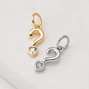 Op de afbeelding: Twee metalen vraagtekenbedels. Eén is goudkleurig, de andere zilverkleurig. Elke bedel heeft een hartvormige edelsteen aan de onderkant. De bedels zijn klein en waarschijnlijk bedoeld voor het maken van sieraden of als hangers.