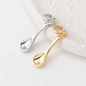 Può includere: Due ciondoli a forma di cucchiaio, uno dorato e uno argentato, appesi ad anelli a salto. I cucchiai sono piccoli e hanno una ciotola arrotondata.