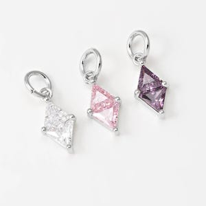 Puede incluir: Tres colgantes de piedras preciosas con forma de diamante. Un colgante tiene una piedra transparente, otro una piedra rosa y el tercero una piedra morada. Cada colgante está unido a un lazo plateado.