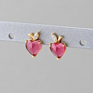 May include: A pair of heart-shaped earrings with a gold-colored setting. The earrings feature a faceted, deep pink gemstone in a heart shape, with a small leaf-like accent at the top. The earrings are displayed on a silver bar.