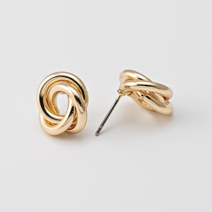 May include: A pair of gold-colored stud earrings. Each earring features a knotted design, with multiple loops intertwined to create a textured appearance. The earrings have a post and backing for secure wear. The background is a plain white.