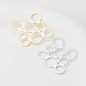 Peut inclure: Deux breloques ajourées en métal. L'une est de couleur or, l'autre de couleur argent. Chaque breloque présente un groupe de cercles interconnectés, créant un design abstrait et bulleux. Les breloques sont présentées sur une surface blanche.