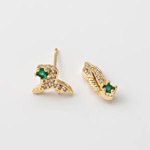 Peut inclure: Une paire de boucles d'oreilles dorées. Chaque boucle d'oreille présente un motif orné de petites pierres précieuses claires et vertes. Les boucles d'oreilles sont posées sur un fond blanc.