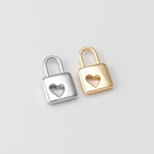 May include: Two small, square padlocks with heart-shaped cutouts. One is silver-colored, the other is gold-colored. The padlocks have a simple, classic design, and are likely intended for jewelry or craft projects.