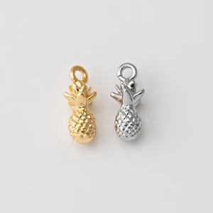 May include: Two small pineapple charms, one gold-colored and one silver-colored, are displayed against a white background. The charms have a textured surface and a loop at the top for attaching to jewelry. These charms are ideal for crafting.