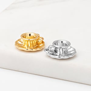 Può includere: Due tazze da tè in miniatura con piattini. Una è color oro, l'altra color argento. Entrambe hanno un bordo smerlato. Le tazze da tè sono posizionate su una superficie bianca.