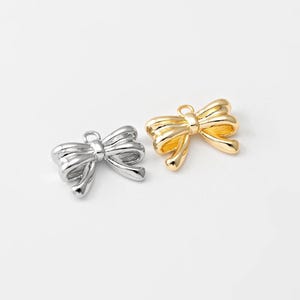 Op de afbeelding: Twee metalen strikcharms, een gouden en een zilveren. De strikken zijn gedetailleerd en hebben een driedimensionale uitstraling.