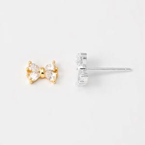 Pode incluir: Um par de brincos de pressão de ouro e prata com um pequeno design de laço com pedras de zircônia cúbica transparentes.