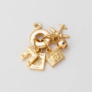 Puede incluir: Pulsera de charms de oro con charms de temática de viaje, incluyendo un pasaporte, un avión, una cámara y el Arco del Triunfo.