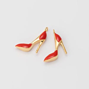 Könnte beinhalten: Zwei rote und goldene High-Heel-Schuh-Anhänger. Die Anhänger haben eine spitze Spitze und einen dünnen Absatz. Der obere Teil des Schuhs ist rot, und die Sohle und der Absatz sind goldfarben. Auf weißem Hintergrund.