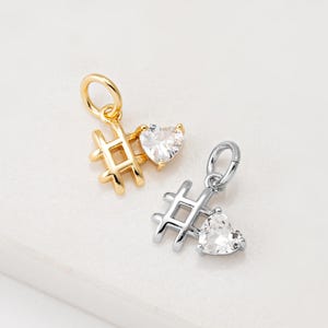 Könnte beinhalten: Zwei kleine Anhänger, einer goldfarben und einer silberfarben, jeweils mit einem Hashtag-Symbol und einem herzförmigen, klaren Edelstein. Die Anhänger haben oben eine kleine Schlaufe zum Befestigen an einer Halskette oder einem Armband.