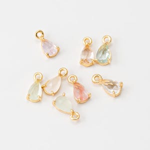 Può includere: Una collezione di ciondoli a forma di goccia di pietre preziose in vari colori pastello, tra cui rosa, verde e trasparente. Ogni ciondolo ha una montatura in metallo color oro e un piccolo anello nella parte superiore per il fissaggio ai gioielli. I ciondoli sono su una superficie bianca.