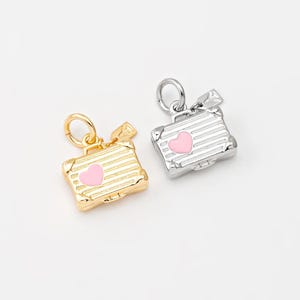 Könnte beinhalten: Zwei kleine Kofferanhänger, einer goldfarben und einer silberfarben, jeweils mit einem rosa Herzdetail. Die Anhänger haben ein strukturiertes, gestreiftes Design und eine kleine Schlaufe zum Befestigen. Die Anhänger sind klein, etwa 1,3 cm lang.