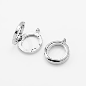 Puede incluir: Dos colgantes de medallón redondos de color plateado con inserciones de vidrio transparente. Un medallón está abierto, revelando el interior, mientras que el otro está cerrado. Ambos tienen pequeños bucles para sujetar a una cadena o cordón.