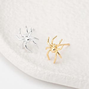 Op de afbeelding: Een paar spinnenoorbellen, een zilveren en een gouden. De oorringen zijn klein en delicaat en zouden een geweldig cadeau zijn voor iedereen die van spinnen houdt.