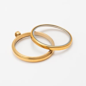 Puede incluir: Dos marcos de colgante circulares dorados con inserciones de vidrio transparente. Un marco se superpone ligeramente al otro. Los marcos tienen un acabado metálico brillante y un pequeño lazo para colgar. El fondo es blanco liso.