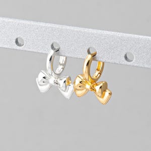 Puede incluir: Dos pares de pendientes de aro con un dije de lazo. Un par es dorado y el otro es plateado.