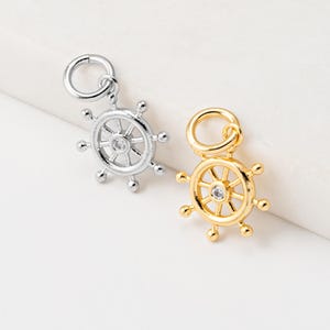 May include: Two nautical-themed charms in silver and gold. Each charm is shaped like a ship's wheel with a central gemstone. The silver charm has a bright, polished finish, while the gold charm has a warm, shiny appearance. Both have a loop for attaching to jewelry.