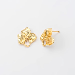 Puede incluir: Un par de pendientes de oro. Cada pendiente presenta un grupo de tres hojas detalladas. Los pendientes están sobre un fondo blanco, resaltando el diseño intrincado y el acabado metálico.