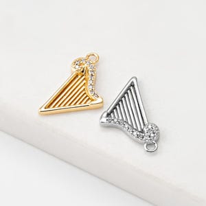 May include: Two harp-shaped charms, one gold-toned and one silver-toned, each with a curved, embellished top and vertical lines. The gold charm has a warm, shiny finish, while the silver charm has a cool, metallic appearance. Both charms have a small loop for attachment.