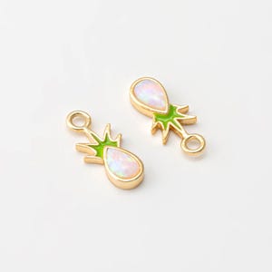 May include: Two gold-colored charm pendants in the shape of a pineapple. Each pendant features a pear-shaped opal-like stone, green enamel accents, and a small loop for attachment. The pendants are on a white background.