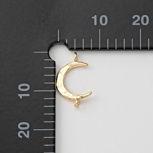 Könnte beinhalten: Ein goldfarbener Halbmond-Anhänger mit strukturierter Oberfläche. Der Anhänger hat oben eine kleine Schlaufe zum Aufhängen und unten eine kleine Verlängerung. Der Anhänger befindet sich vor einem schwarz-weißen Hintergrund mit Messmarkierungen.