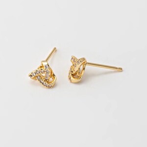 May include: A pair of gold-coloured stud earrings. Each earring has a knot design, encrusted with small, clear gemstones. The earrings are set against a plain white background, highlighting their intricate details and sparkle.