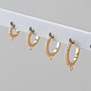 Pode incluir: Quatro brincos de argola dourados com uma fileira de pedras de zircônia cúbica transparentes. Cada brinco tem um aro de junção para prender um berloque ou um pingente.