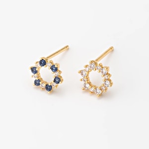 Może przedstawiać: Para złotych kolczyków sztyftów. Jeden kolczyk ma okrągły wzór z naprzemiennymi niebieskimi i przezroczystymi kamieniami. Drugi kolczyk ma podobny wzór, ale tylko z przezroczystymi kamieniami. Kolczyki są na białym tle.