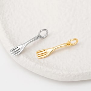 May include: Two small fork charms, one silver-colored and one gold-colored, are displayed on a white surface. Each charm has a loop at the top for attaching to a necklace or bracelet. The forks have three tines.