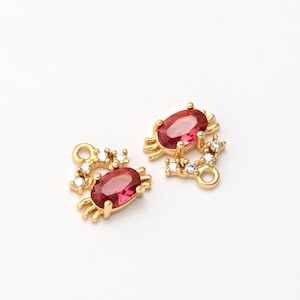 May include: Two gold-colored jewelry charms. Each charm features an oval, ruby-colored stone at the center, surrounded by smaller, clear stones. The charms have a crab-like design with small, curved legs and a loop for attachment. The background is white.
