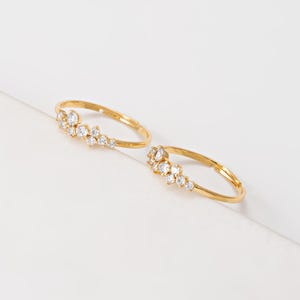 May include: Two gold-colored rings with a cluster of small, clear gemstones. The gemstones are arranged in a curved pattern along the top of the ring band. The rings are set against a white background.