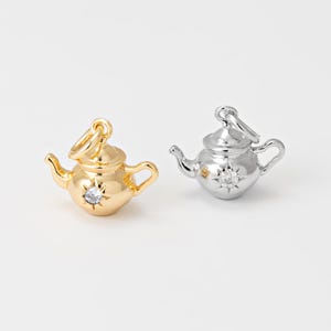 May include: Two small teapot charms, one gold-toned and one silver-toned, each with a starburst design and a clear gemstone. The charms have a loop at the top for attaching to a necklace or bracelet. The background is plain white.