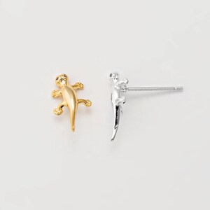 May include: A pair of lizard-shaped stud earrings. One earring is gold-colored, and the other is silver-colored. The earrings are small and detailed, with the lizard's features clearly visible.
