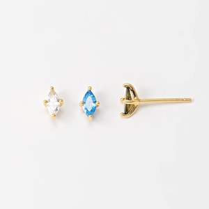 May include: Gold-colored stud earrings with marquise-cut gemstones. One earring features a clear gemstone, the other a blue gemstone. The third earring is a post with a decorative setting.