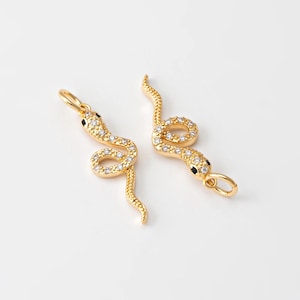 Peut inclure: Deux breloques dorées en forme de serpent, ornées de cristaux transparents et d'yeux noirs. Les breloques serpentines sont enroulées, avec une boucle en haut pour les suspendre. Les breloques sont posées sur un fond blanc.
